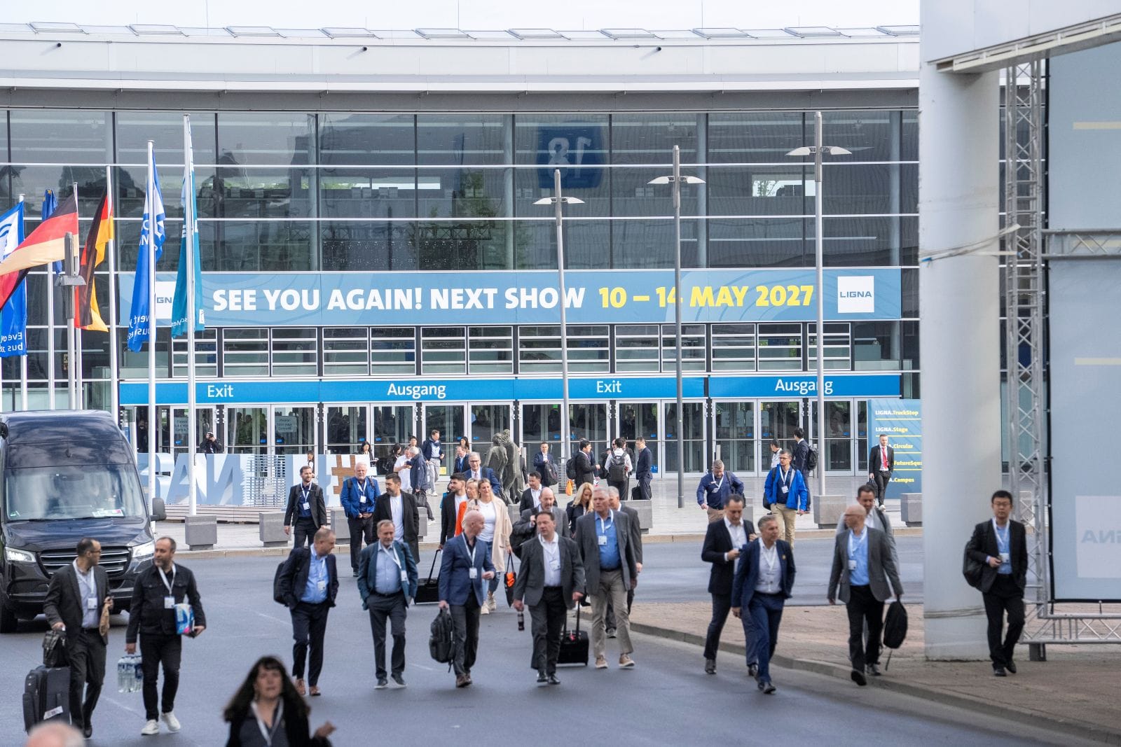 A large group of visitors gathers in front of the exhibition center entrance beneath a prominent yellow sign reading “Save the Date – LIGNA.27, 10–14 May 2027, Exhibition Center Hannover.” The attendees are entering the venue through the marked entrances.